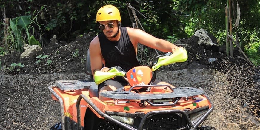 A rider in a helmet and sunglasses steering a quad across the muddy jungle track