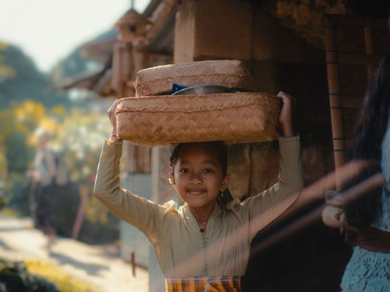 A Balinese girl in a kebaya and sash, smiling as she carries woven offering baskets on her head