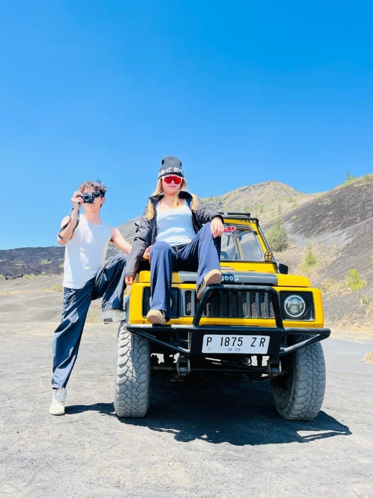 A couple posing with a yellow jeep on Batur’s black sand under a blue sky