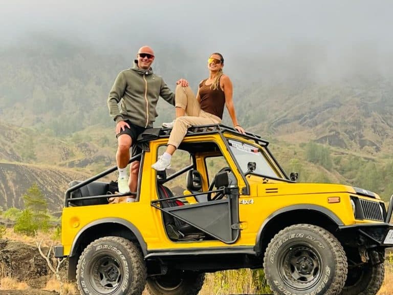 A couple on top of a yellow 4×4 on Batur’s black sand, morning mist on the caldera behind