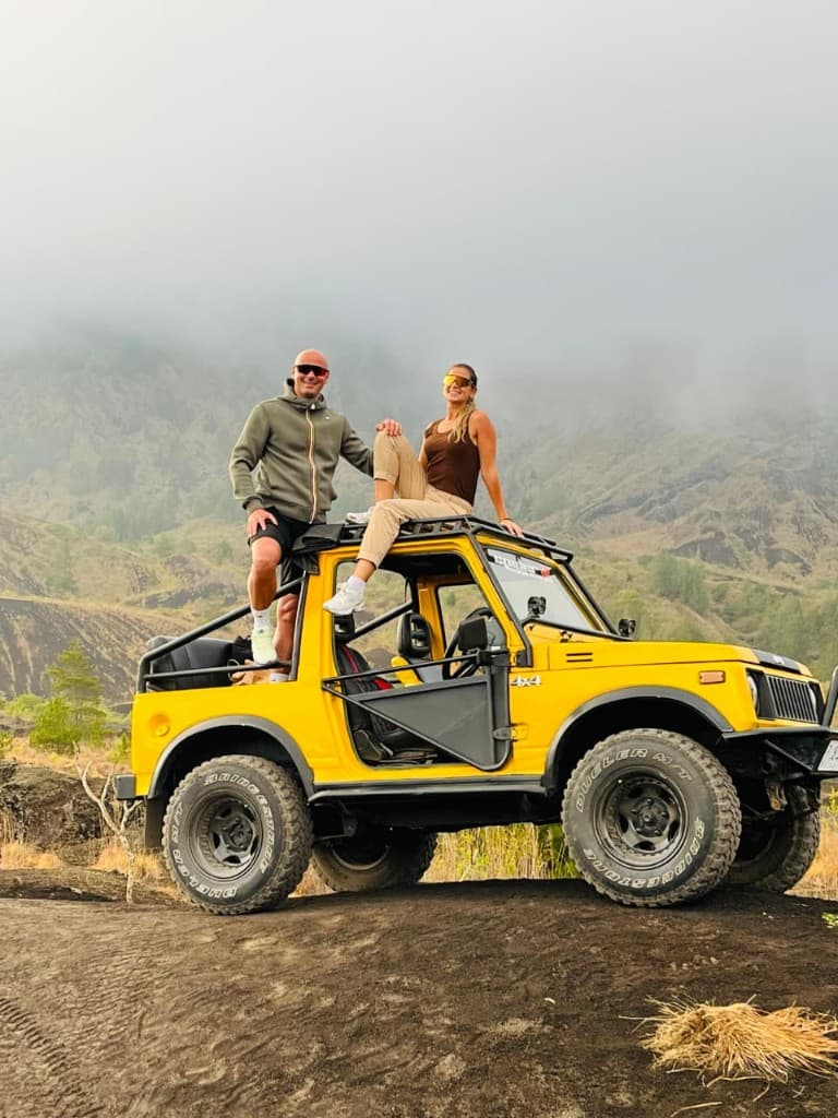 A couple on top of a yellow 4×4 on Batur’s black sand, morning mist on the caldera behind