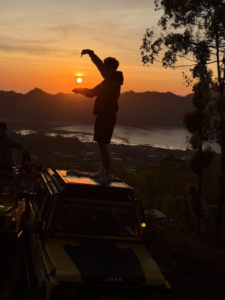 A silhouetted guest on a jeep roof framing the sunrise over Lake Batur
