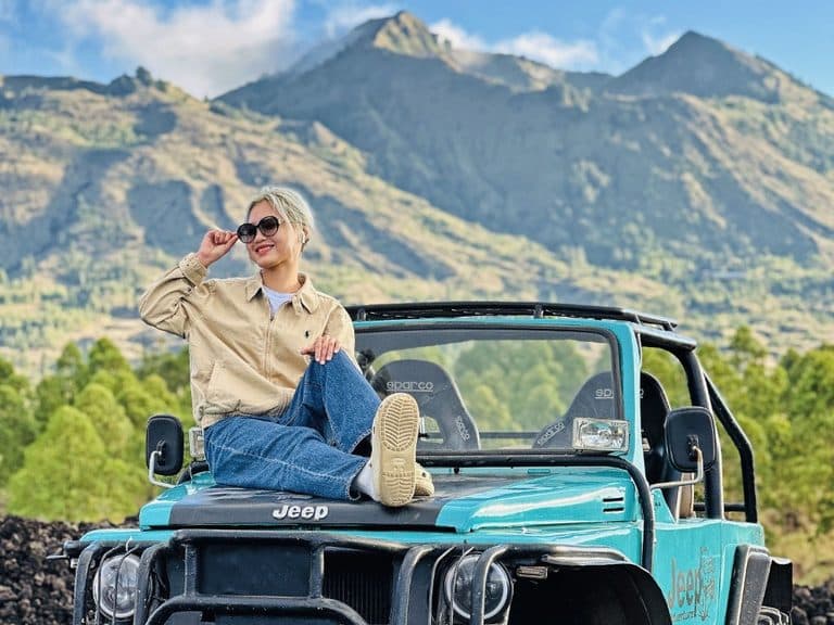 A guest sitting on the bonnet of a teal jeep on the lava field, Mount Batur behind