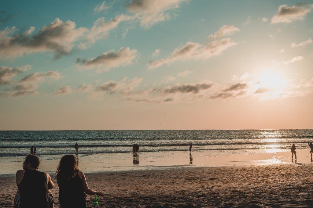Sunset over the sea in Seminyak, two friends sitting on the sand and people wading in the shallows