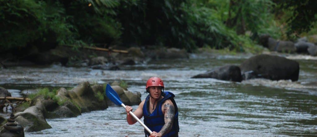 A guest in a red helmet and blue life jacket holding a paddle in the river