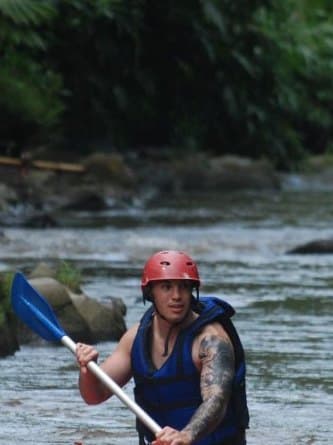 A guest standing in the river in a helmet and life jacket, holding a blue paddle